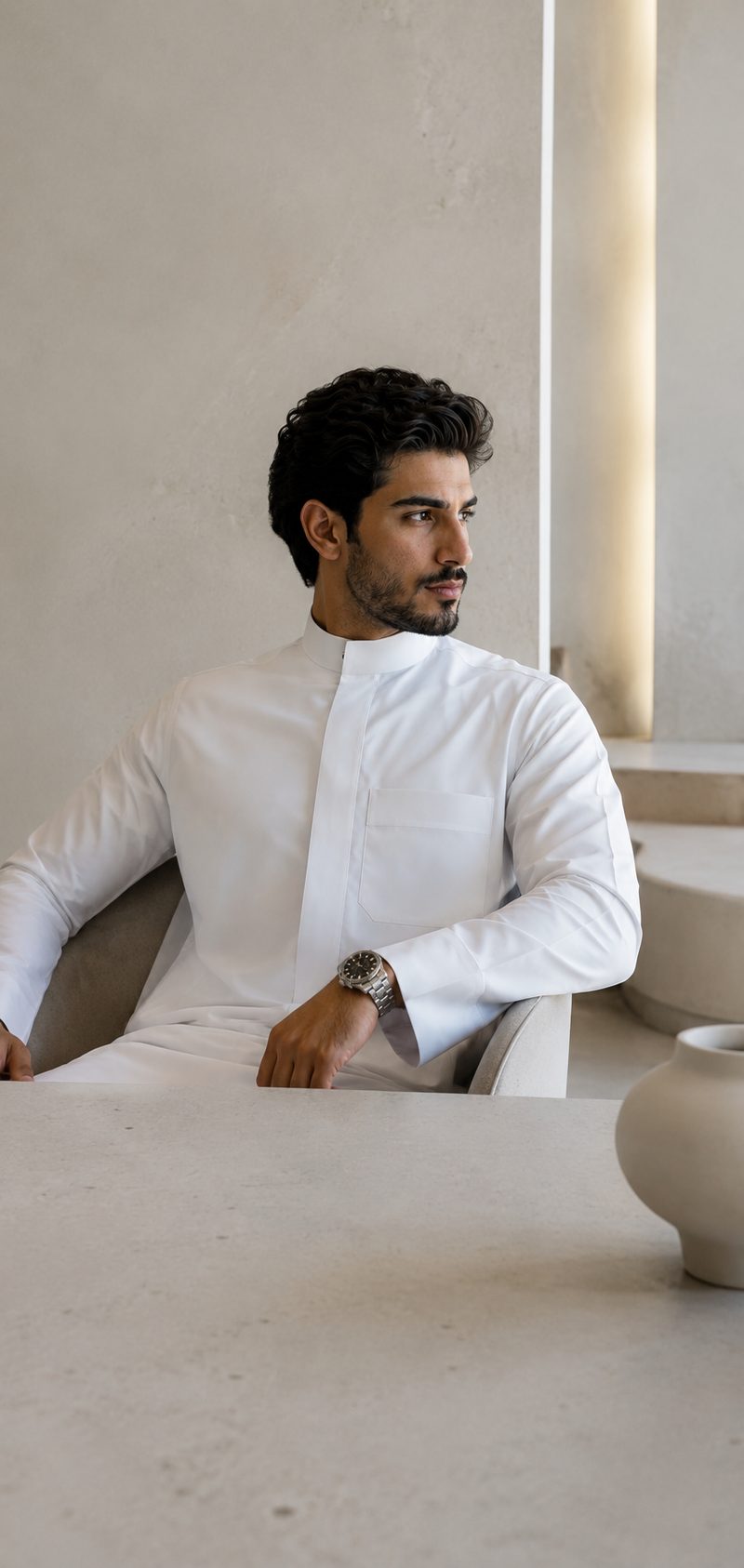 رجل بثوب أبيض يجلس في ردهة هادئة وعلى معصمه ساعة